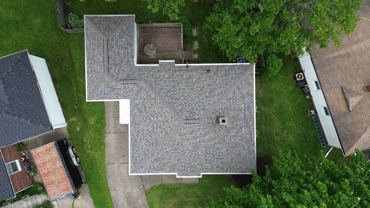 Overhead view of a house with a gray roof, small deck, and green yard, surrounded by trees and other buildings.