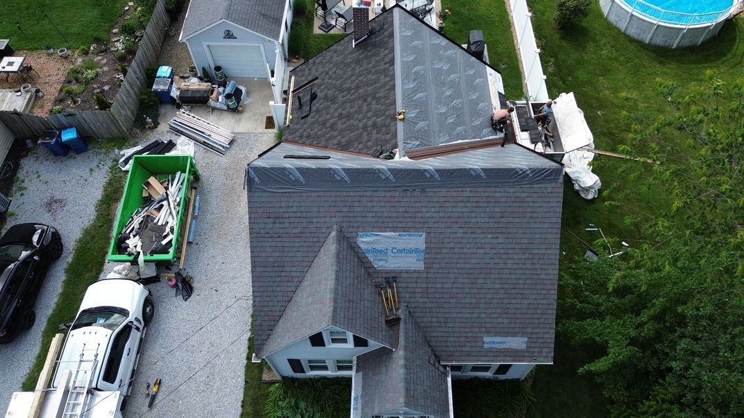 Aerial view of a house with a partially replaced gray roof, construction debris, and a green dumpster in the driveway.