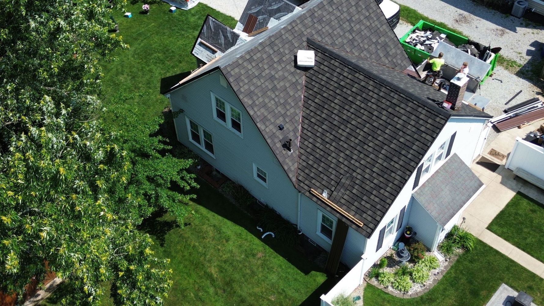 Aerial view of a gray house with a dark roof and green lawn; part of a tree is in the foreground.