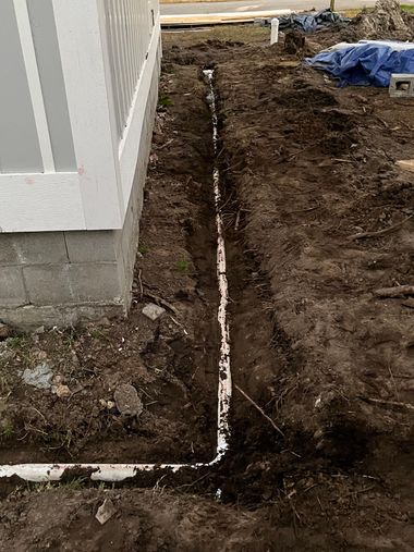 Narrow trench with white PVC pipe running beside a house foundation in a muddy yard.