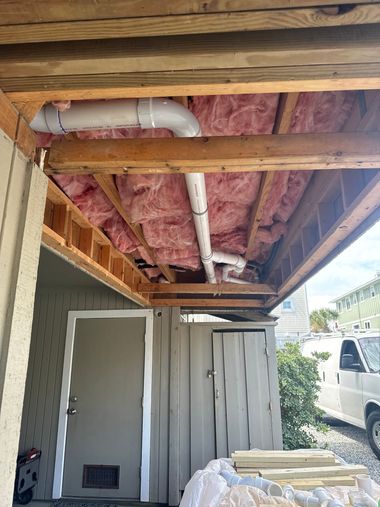 Exposed porch ceiling with pink insulation, white PVC pipes, and stacked lumber beside a shed and white van