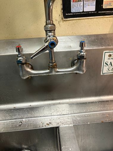 Commercial kitchen sink with a wall-mounted faucet and two blue-handled side valves.