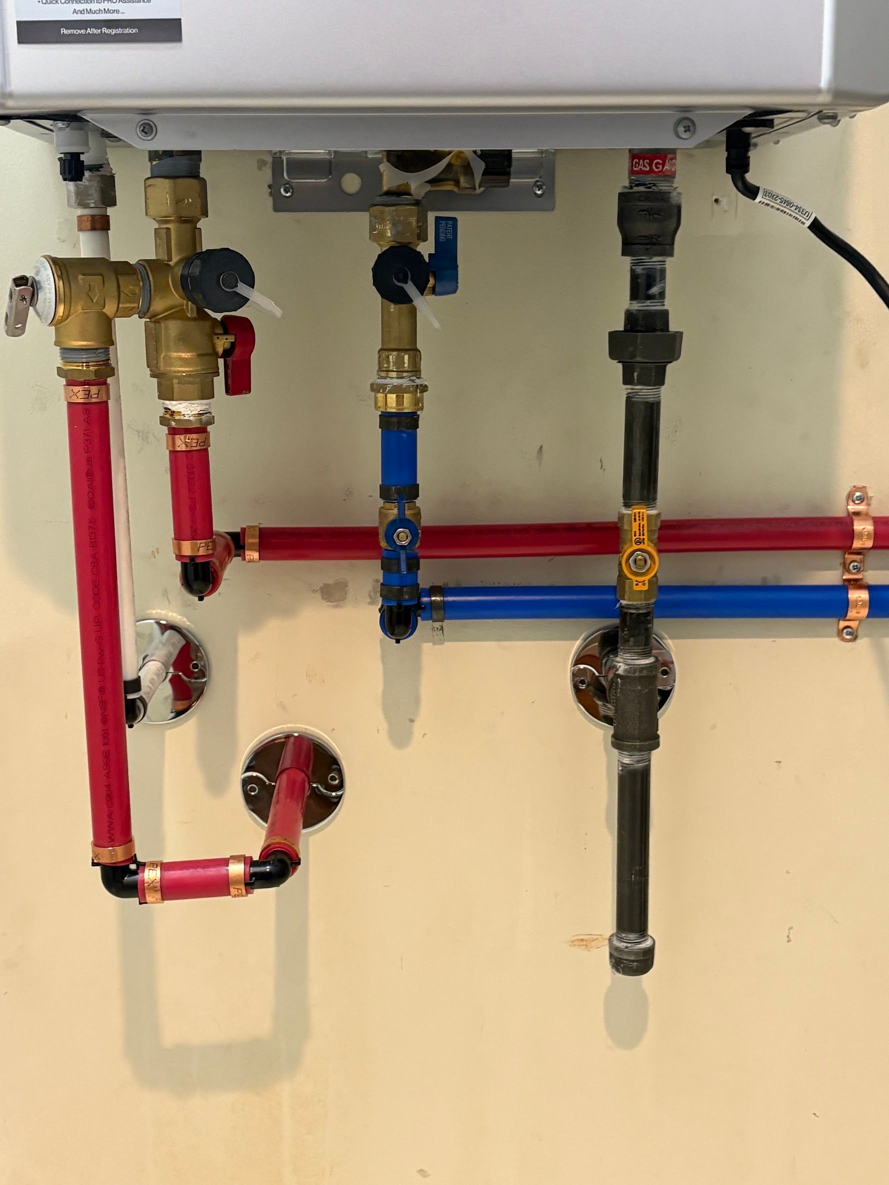 Underside of a boiler with red, blue, and black plumbing pipes and valves against a beige wall