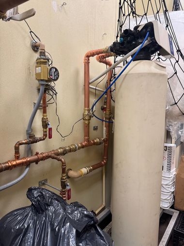 Boiler room with copper pipes, a white water tank, and exposed plumbing on a beige wall.