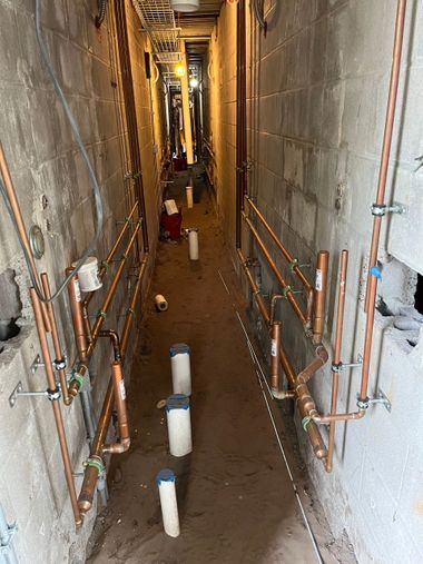 Narrow construction corridor with exposed pipes and wiring, lit by work lights, with workers in the distance