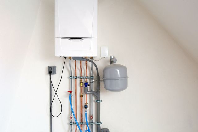 Wall-mounted boiler with exposed pipes, cables, and a gray expansion tank in a white utility room