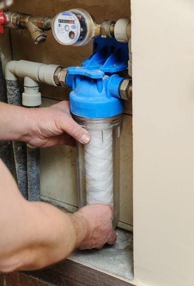Hands installing a blue water filter cartridge under a sink next to a water meter.