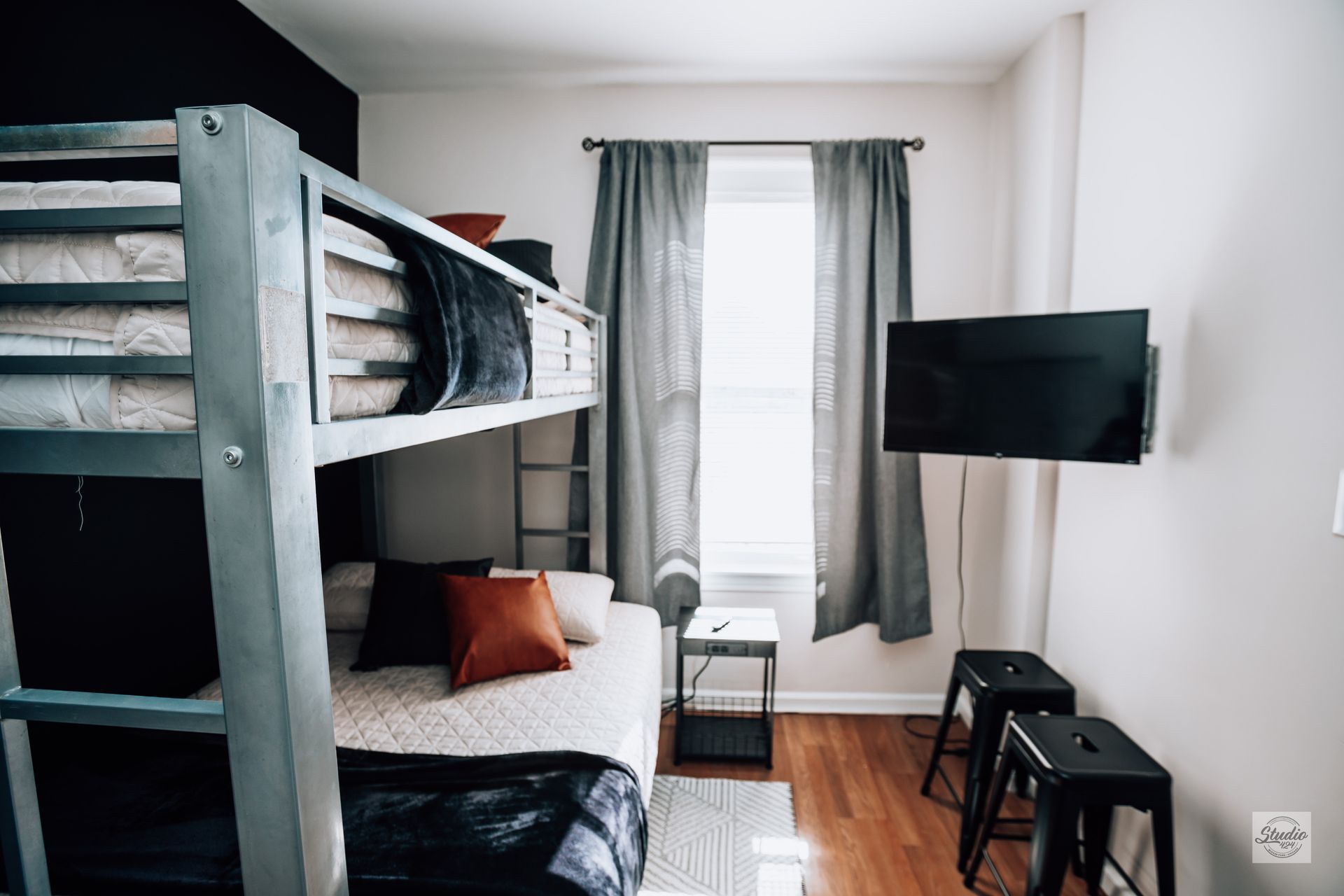 Bunk beds in a room, with a TV, a window with grey curtains, and two black stools.