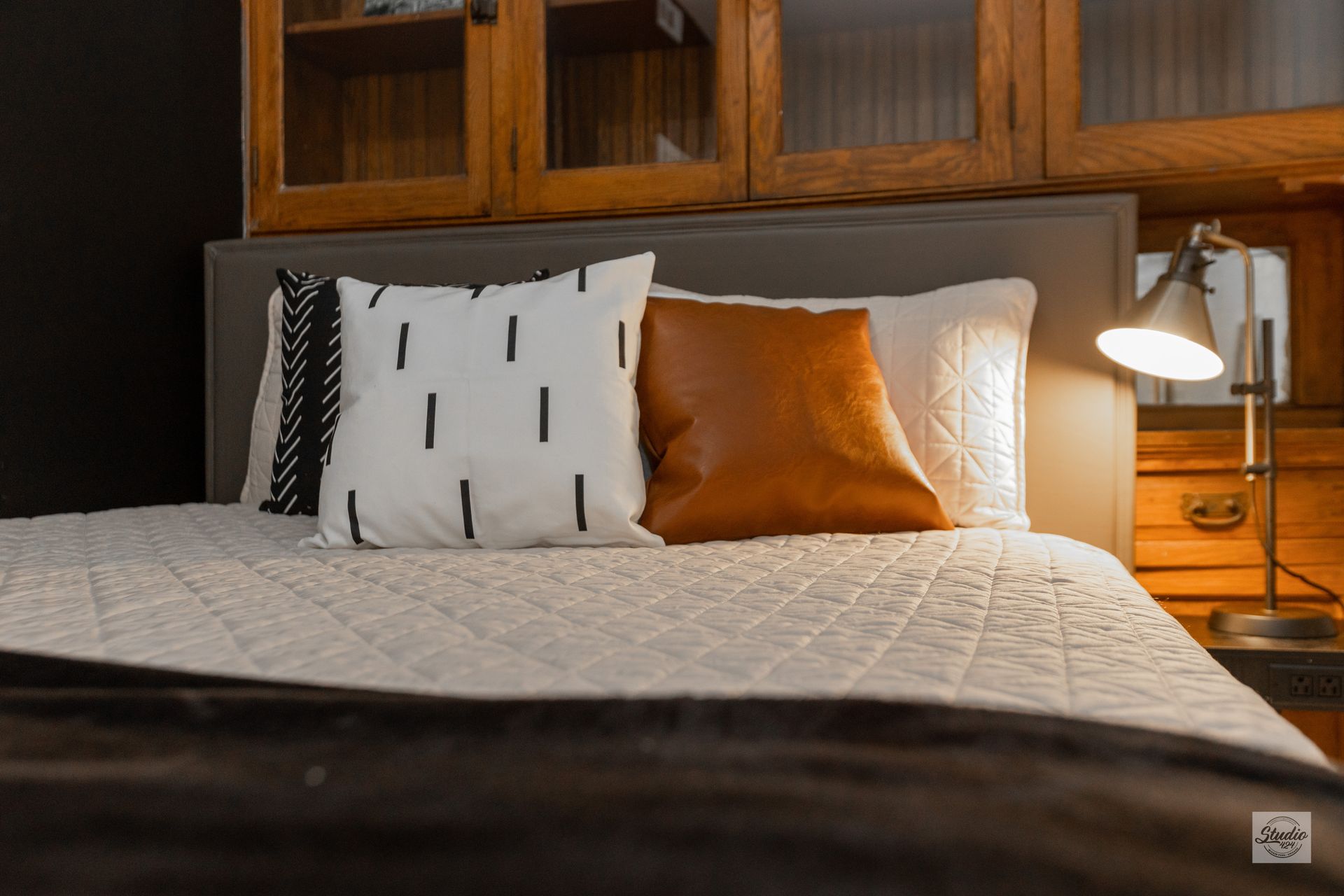 Bed with pillows, headboard, and wooden cabinets above. Lamp on bedside table.