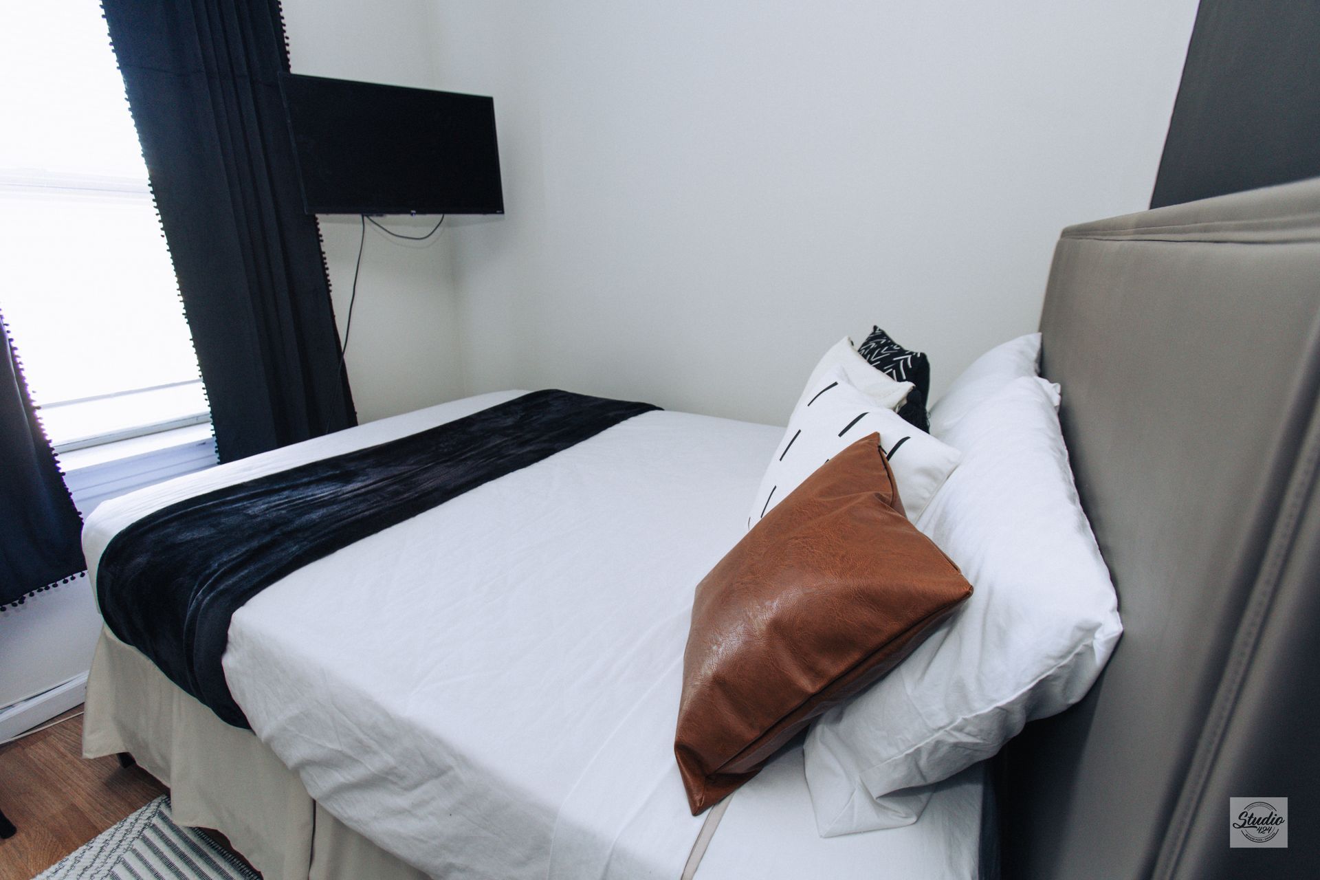 Small bedroom with white bedding, brown and black pillows, a black TV, and a window.