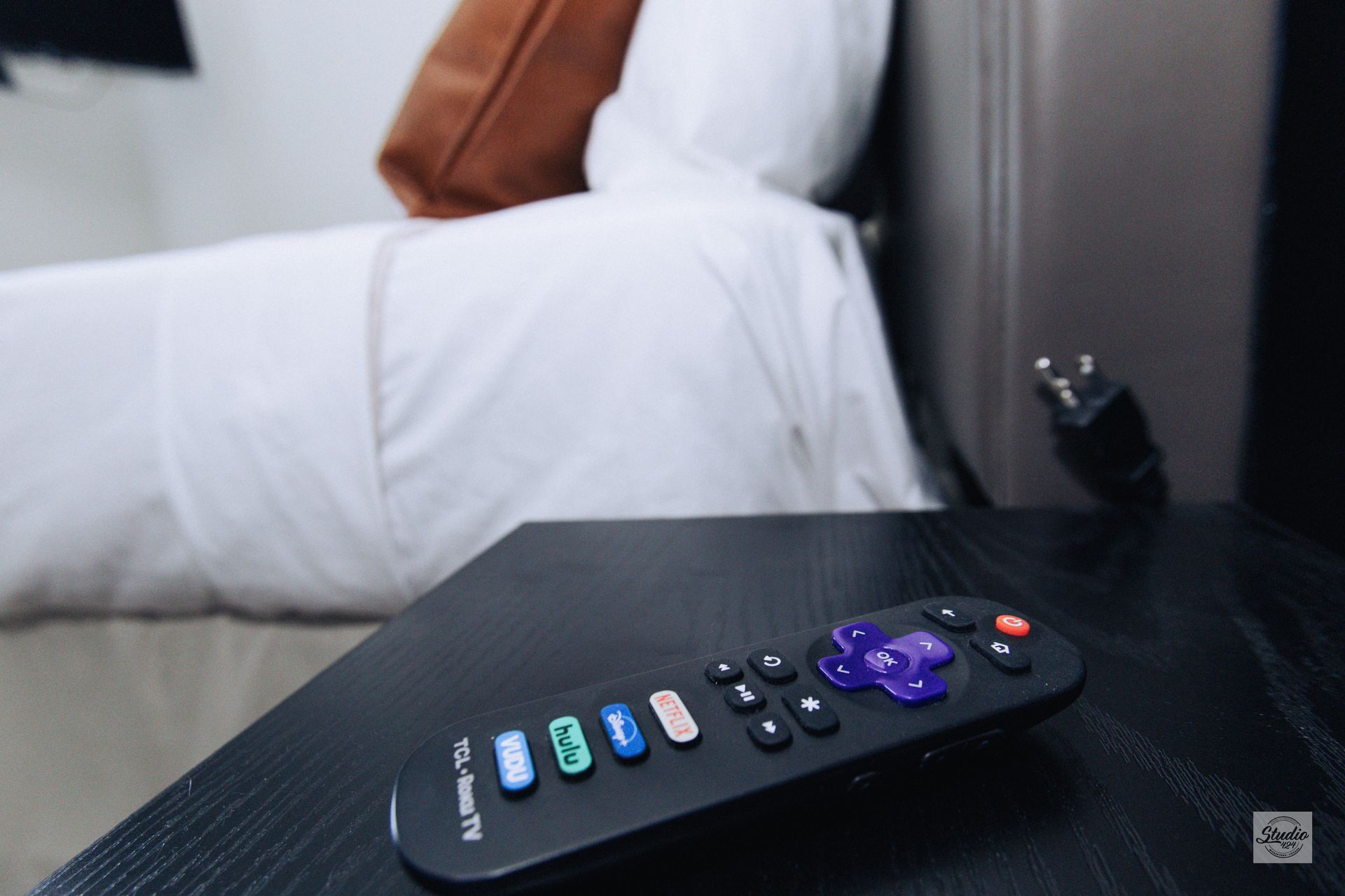 A black Roku remote on a nightstand with a bed in the background.