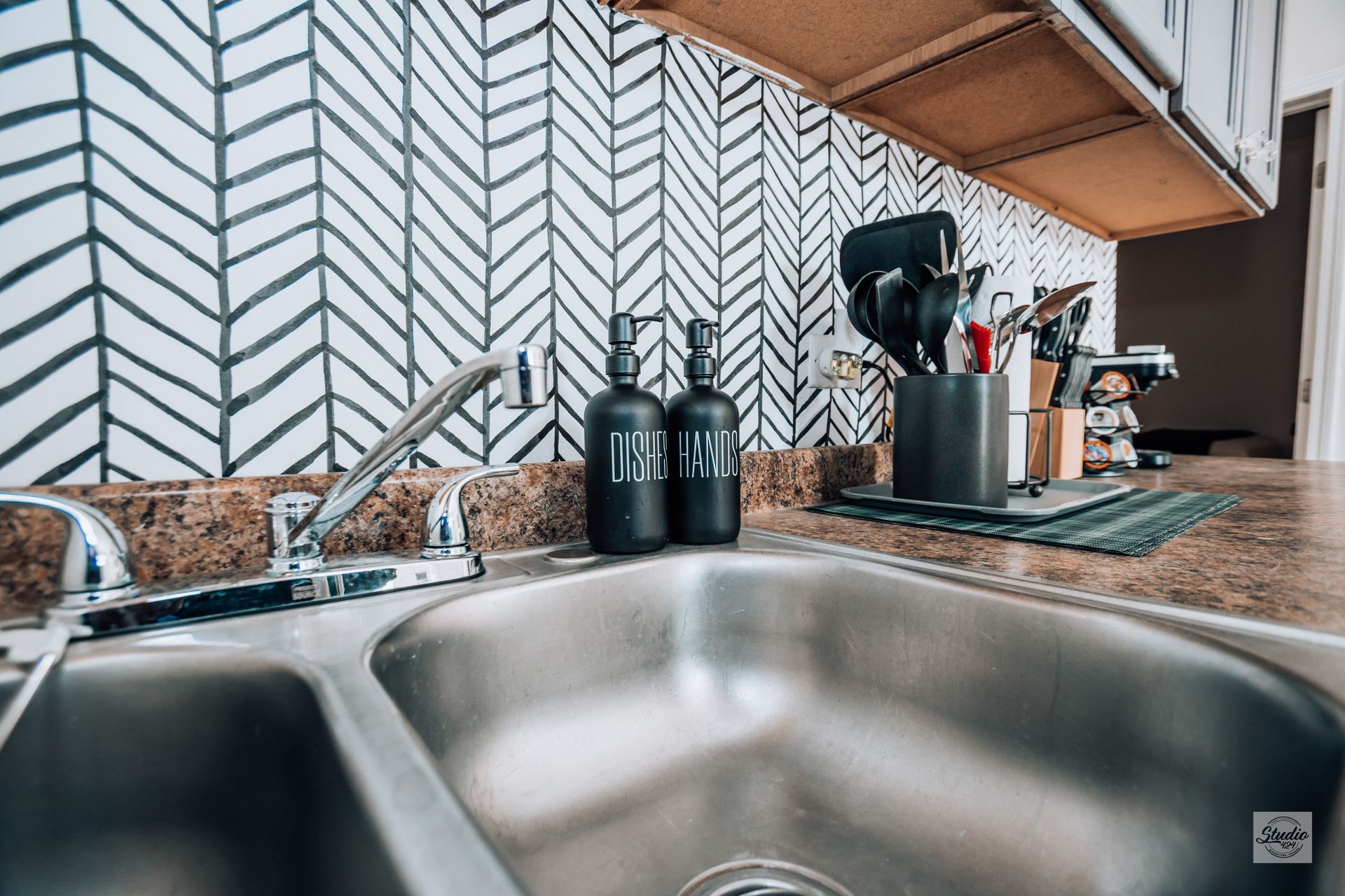 Stainless steel kitchen sink with black soap dispensers, against a patterned backsplash, and utensils.