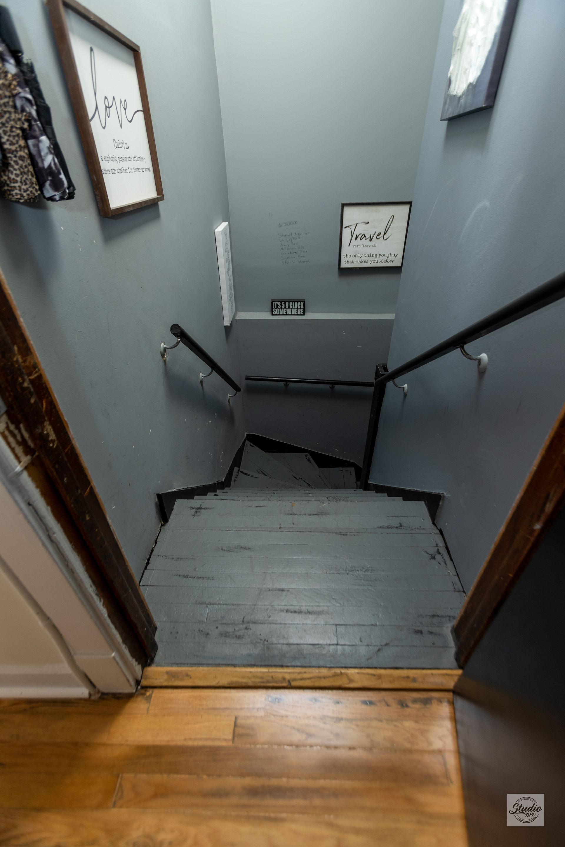 Staircase with gray walls and wooden handrails, leading downwards. Artwork hangs on the walls.