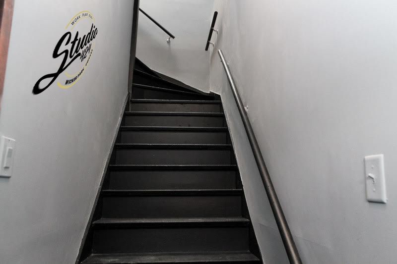 Staircase with black steps and white walls. A metal handrail runs alongside the stairs. A logo is visible on the left wall.