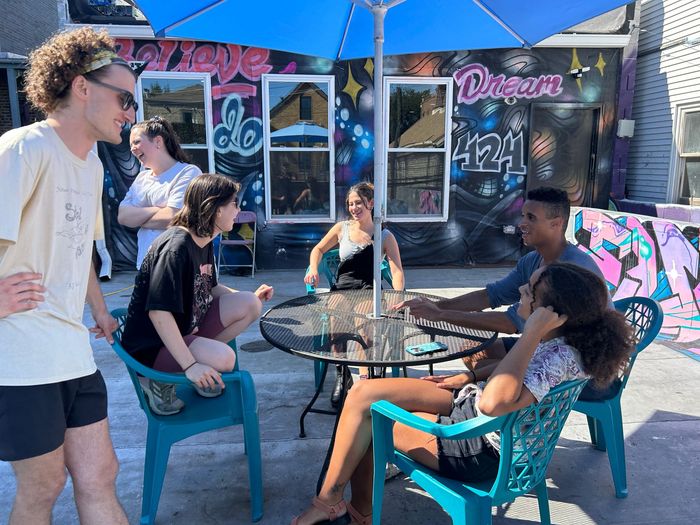 Group of people chatting at an outdoor table under a blue umbrella, colorful graffiti background.
