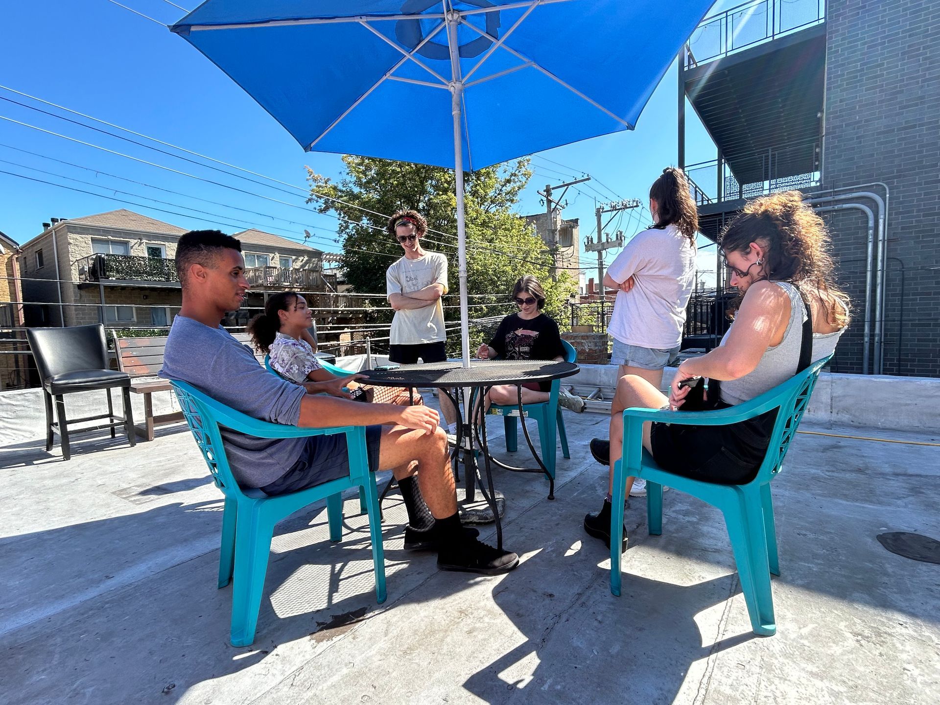 Group of people sitting and standing under a blue umbrella on a patio. Sunny day.