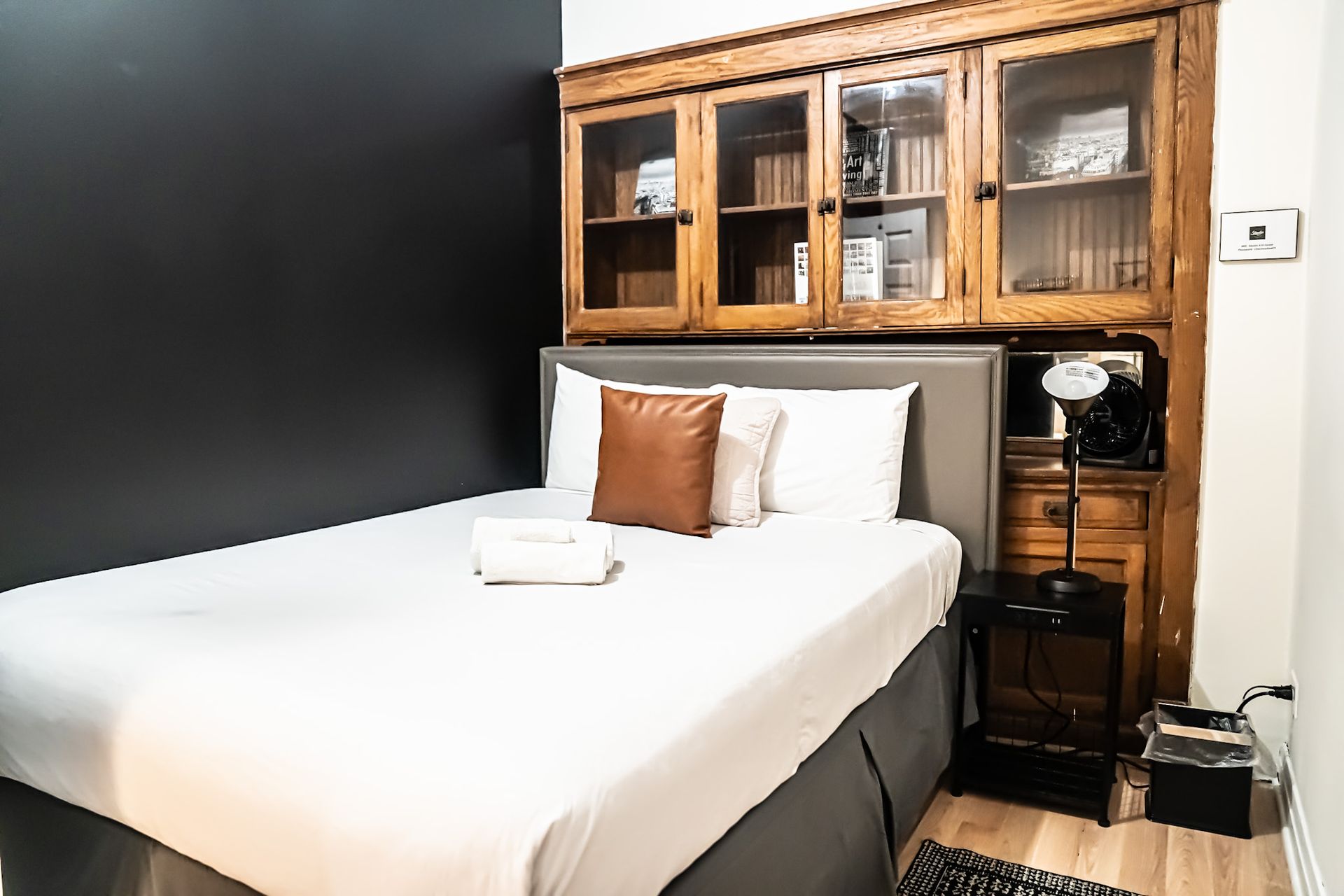 Bedroom with a bed, dark wall, and a wooden cabinet with glass doors; a bedside table and floor heater are also present.