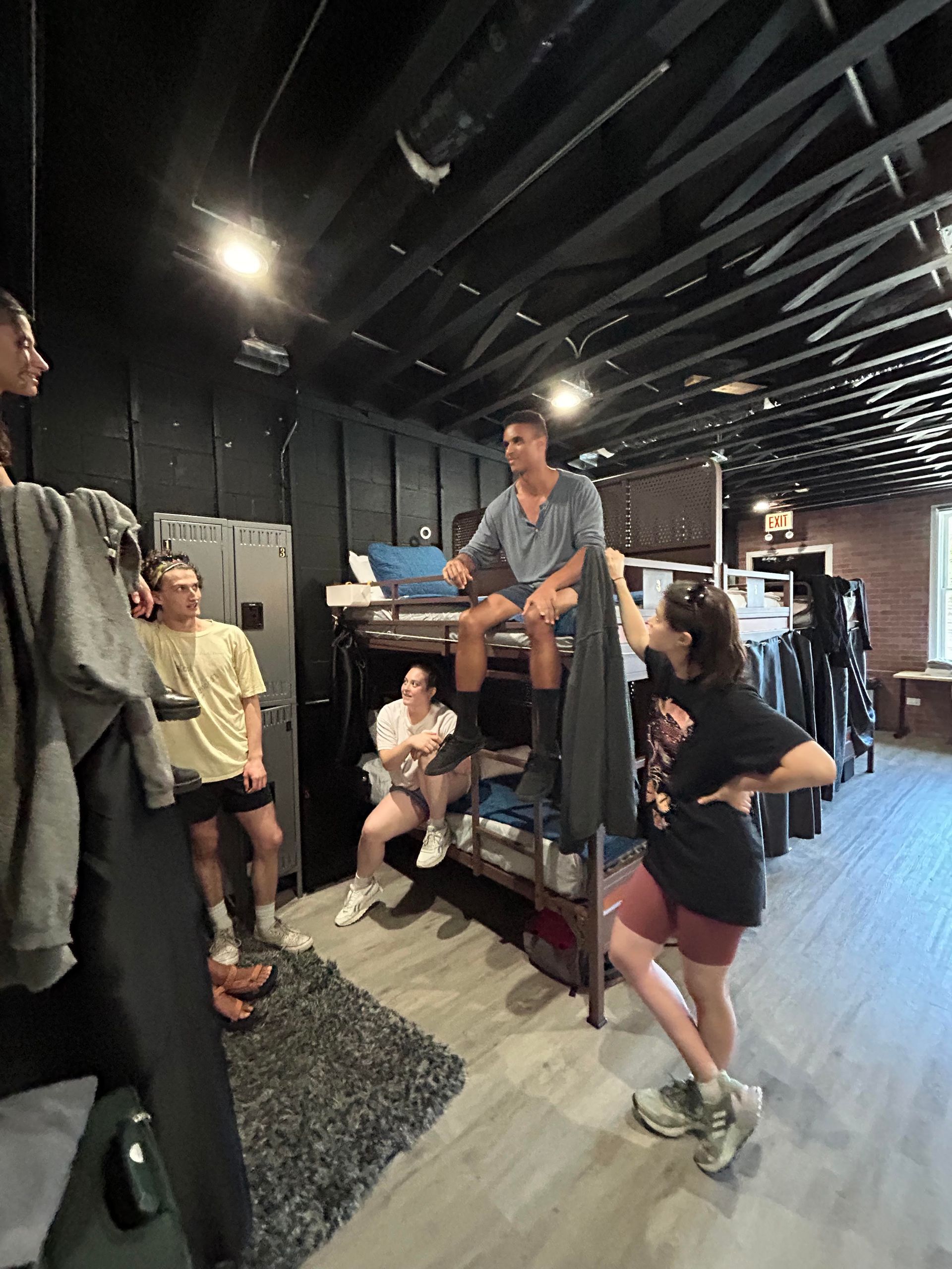 A group of people are gathered in a room with bunk beds. One person sits on the top bunk, interacting with the others.