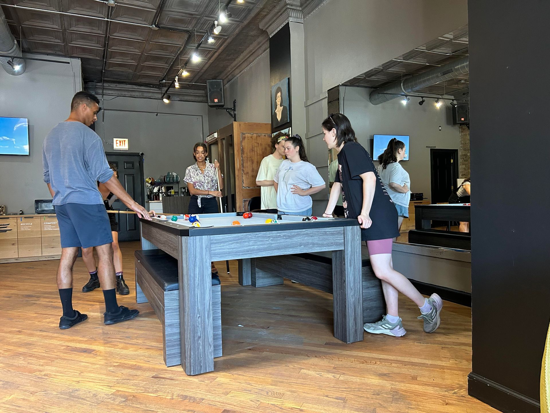 People playing pool in a room with a gray pool table. Various people stand around and are watching the game.