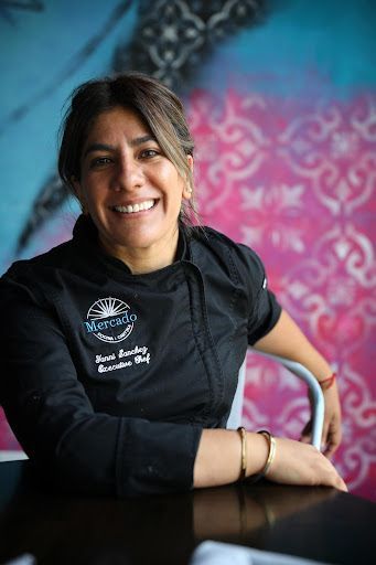 Woman chef smiling, wearing a black jacket with a restaurant logo, seated near a colorful wall.