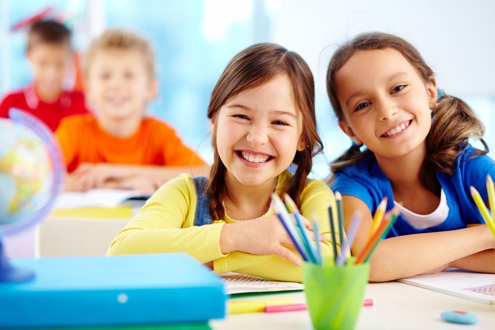 Two Young Girls Are Smiling While Sitting at a Desk in a Classroom — Grandma Rosie’s Quality Long Day Care In Primbee, NSW