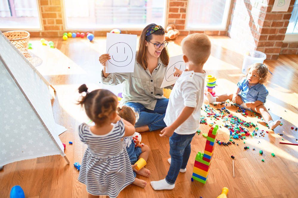 Beautiful Teacher Holding Emotions Emojis With Four Toddlers Around Lots Of Toys At Kindergarten — Grandma Rosie’s Quality Long Day Care In Wollongong, NSW