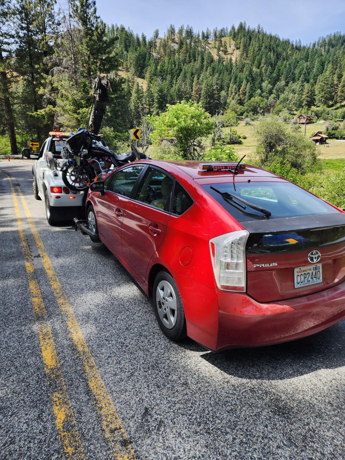 Red Prius towed by a truck on a mountain road beside a forested hillside.