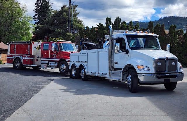 White tow truck pulling a red fire engine on a road with trees and cloudy sky in the background