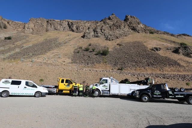 Tow trucks and a white van parked beneath a rocky hillside under a clear blue sky
