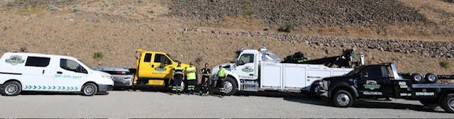 Tow truck and pickup truck wreckers lined up on a dirt roadside beside a white van and yellow service vehicle