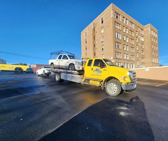 Yellow tow truck carrying a white SUV on a flatbed beside a brick building on a sunny street