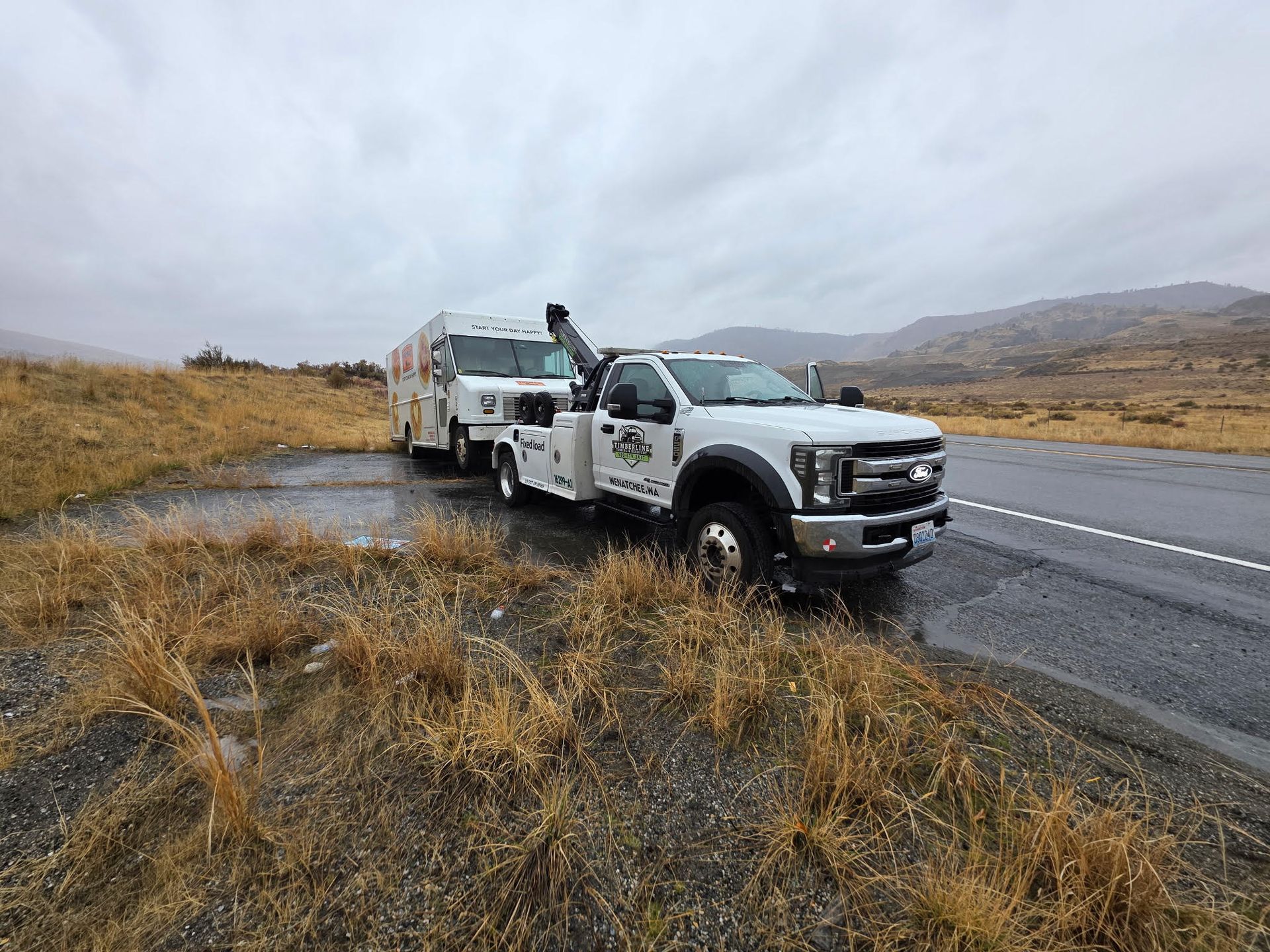 White tow truck on rainy roadside beside a highway in a grassy, mountainous landscape