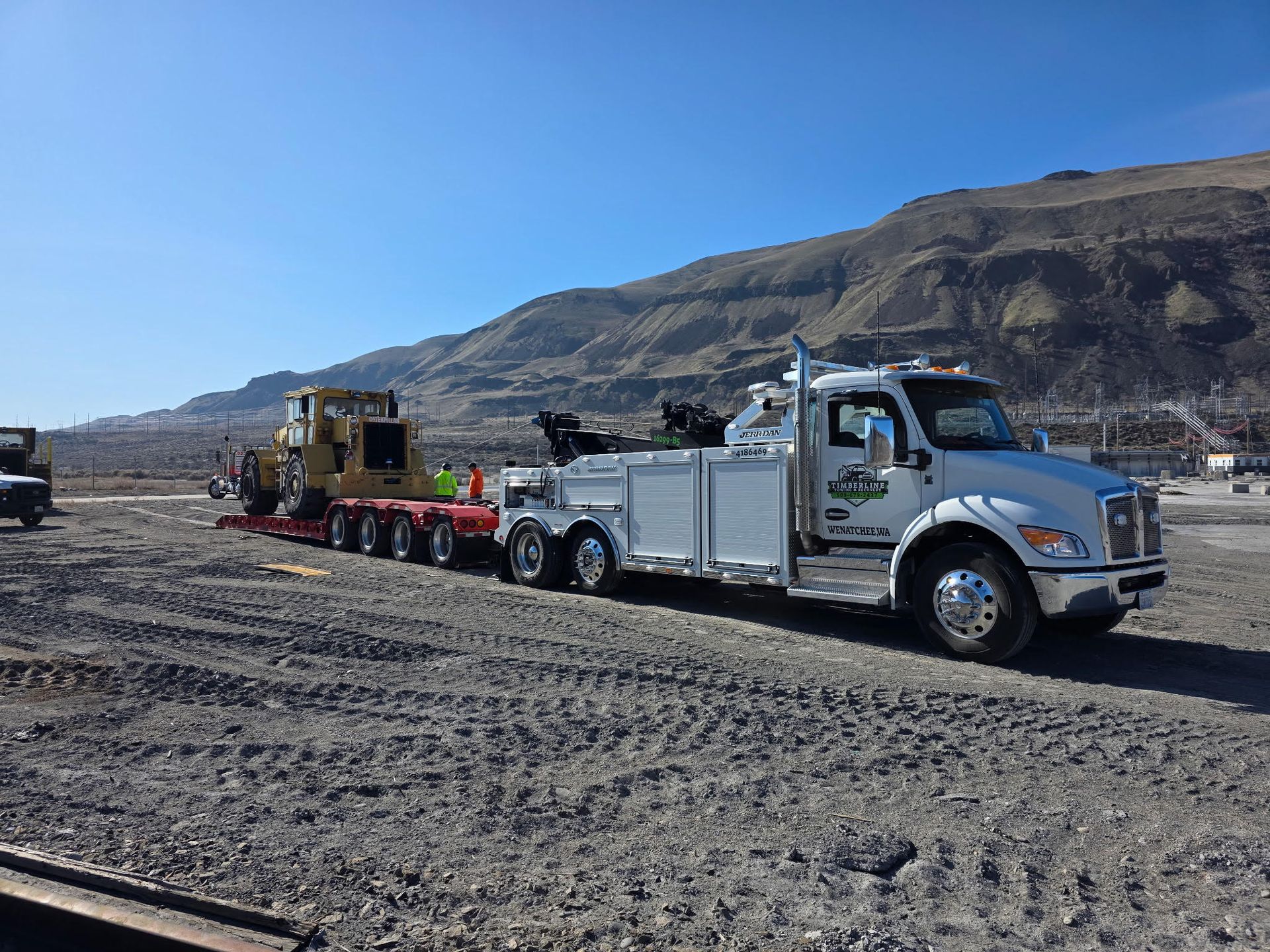 Tow truck hauling heavy equipment across a dirt road in a rocky desert landscape