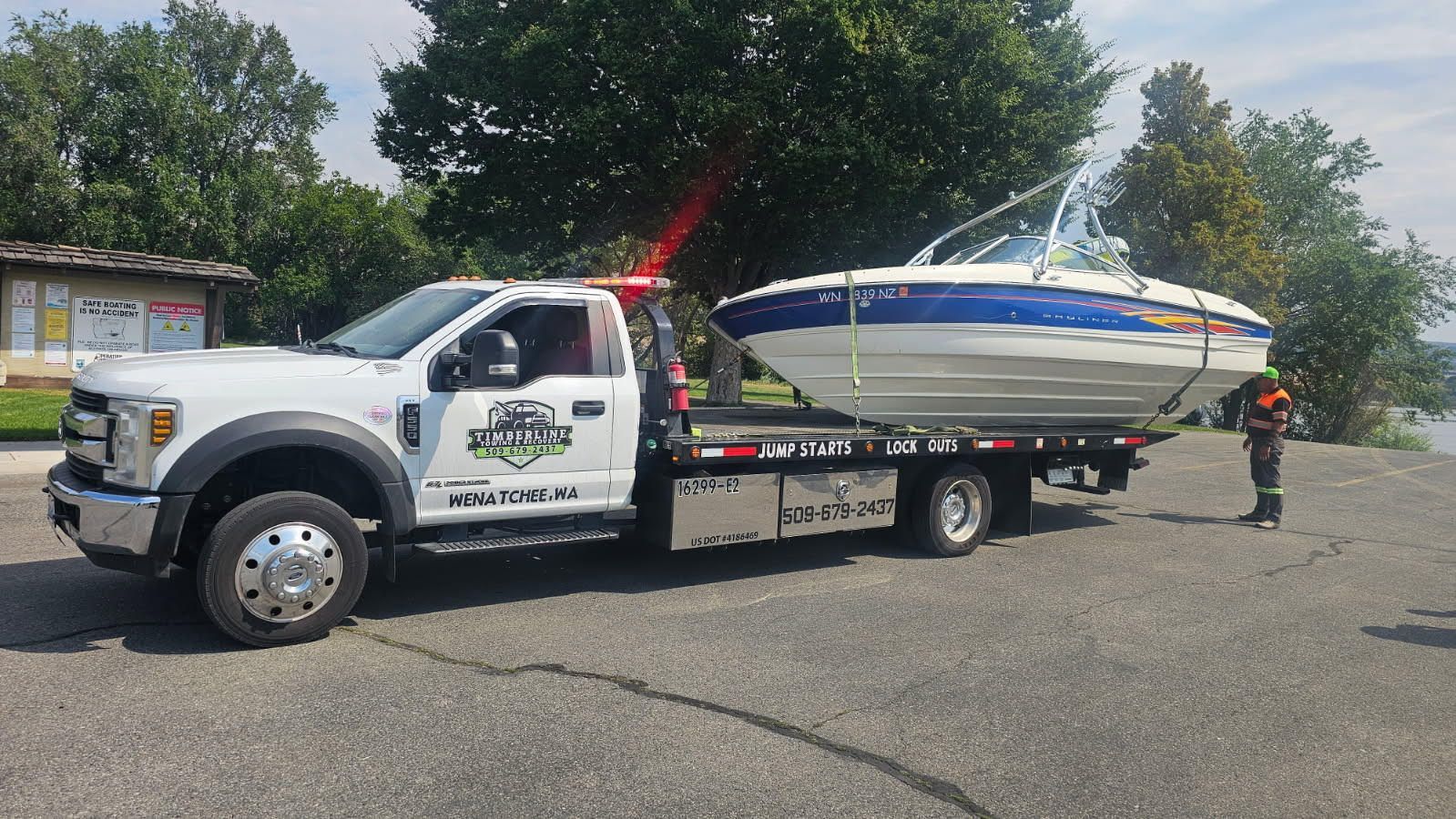 Tow truck carrying a white-and-blue sailboat on a flatbed in a parking lot, with trees in the background