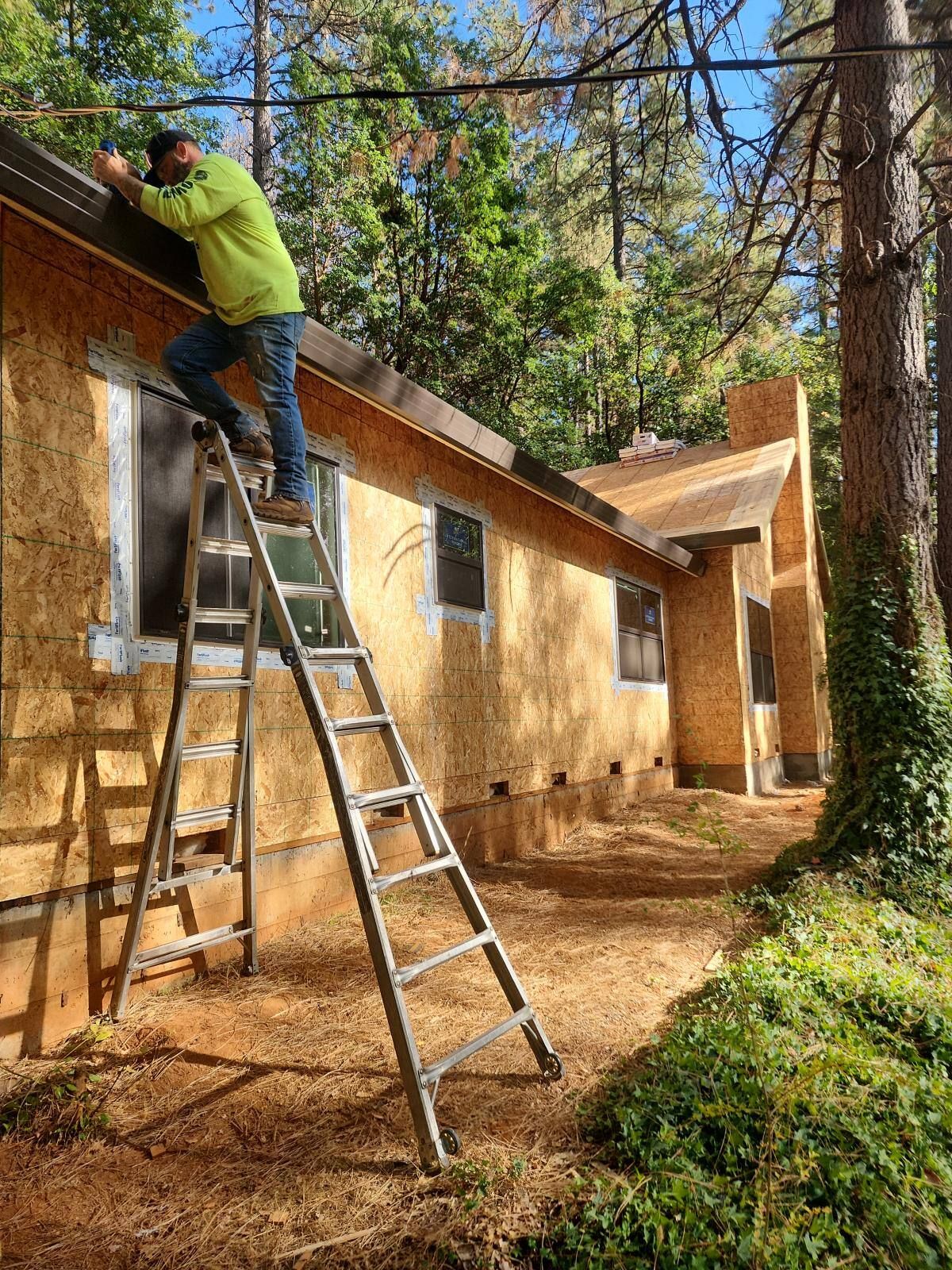 Construction worker on a ladder installing trim on a house with exposed wood siding, in a wooded area.