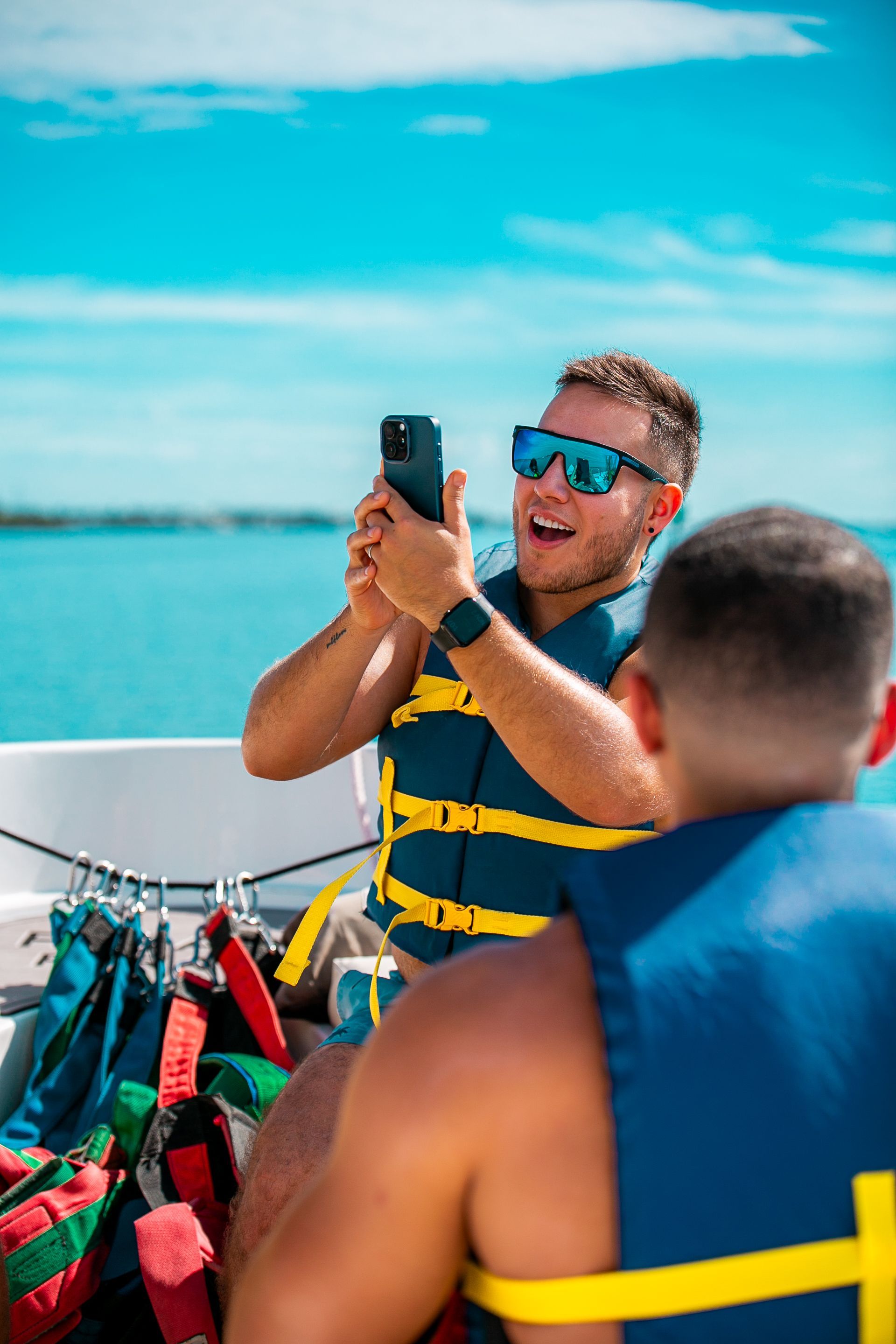 Man on boat takes photo with phone, wearing life vest, blue water, sunny day.