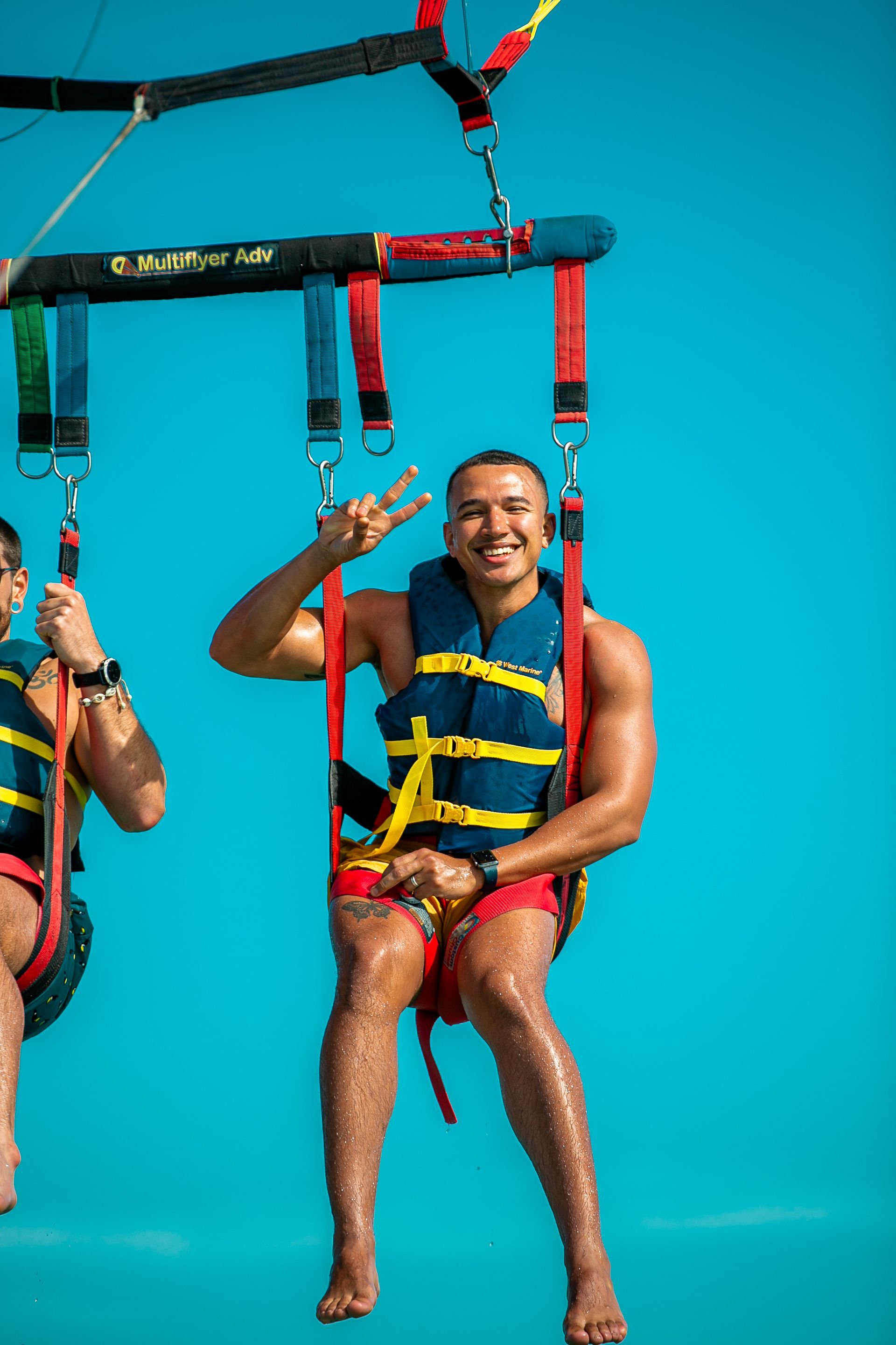 Man parasailing, wearing a life vest, giving the peace sign, against a bright blue sky.