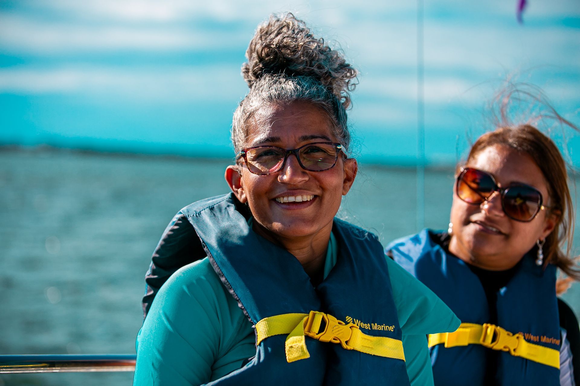 Woman in life vest smiles aboard a boat, another person beside her, ocean background.