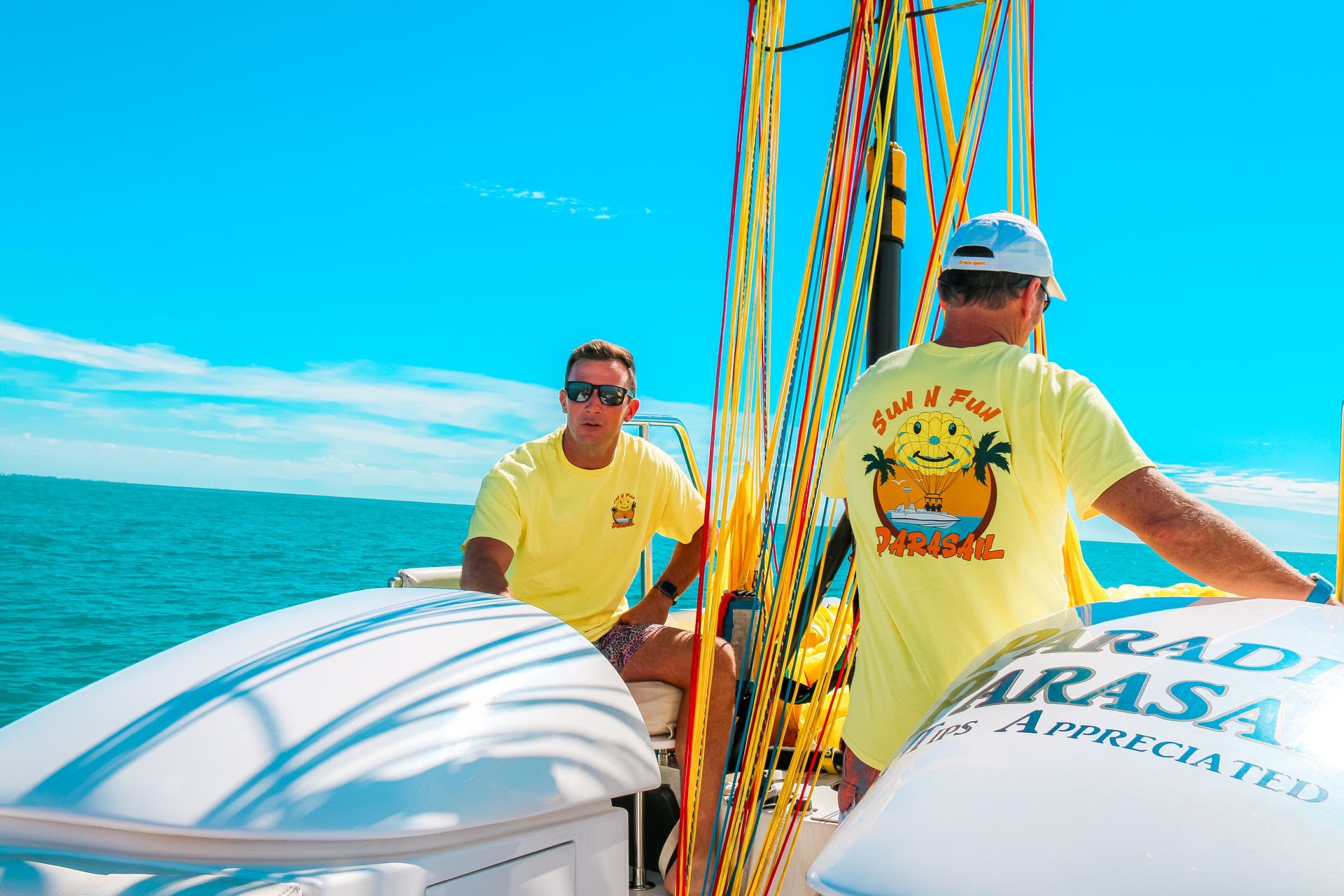 Two men on a boat, bright blue sky, parasailing ropes. One wearing a yellow shirt with a logo on the back.