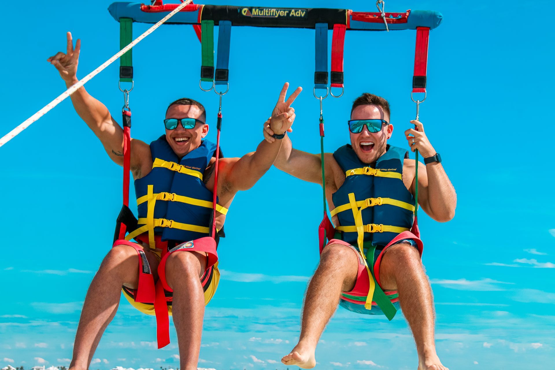 Two people parasailing with blue sky in background. Both wearing sunglasses, life jackets, making peace signs.
