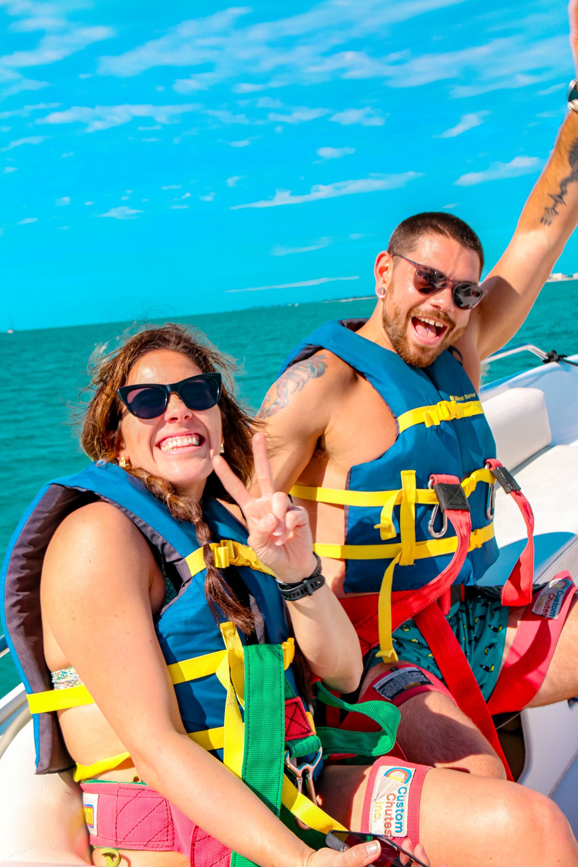 Two people in life vests on a boat, smiling with the ocean behind them.