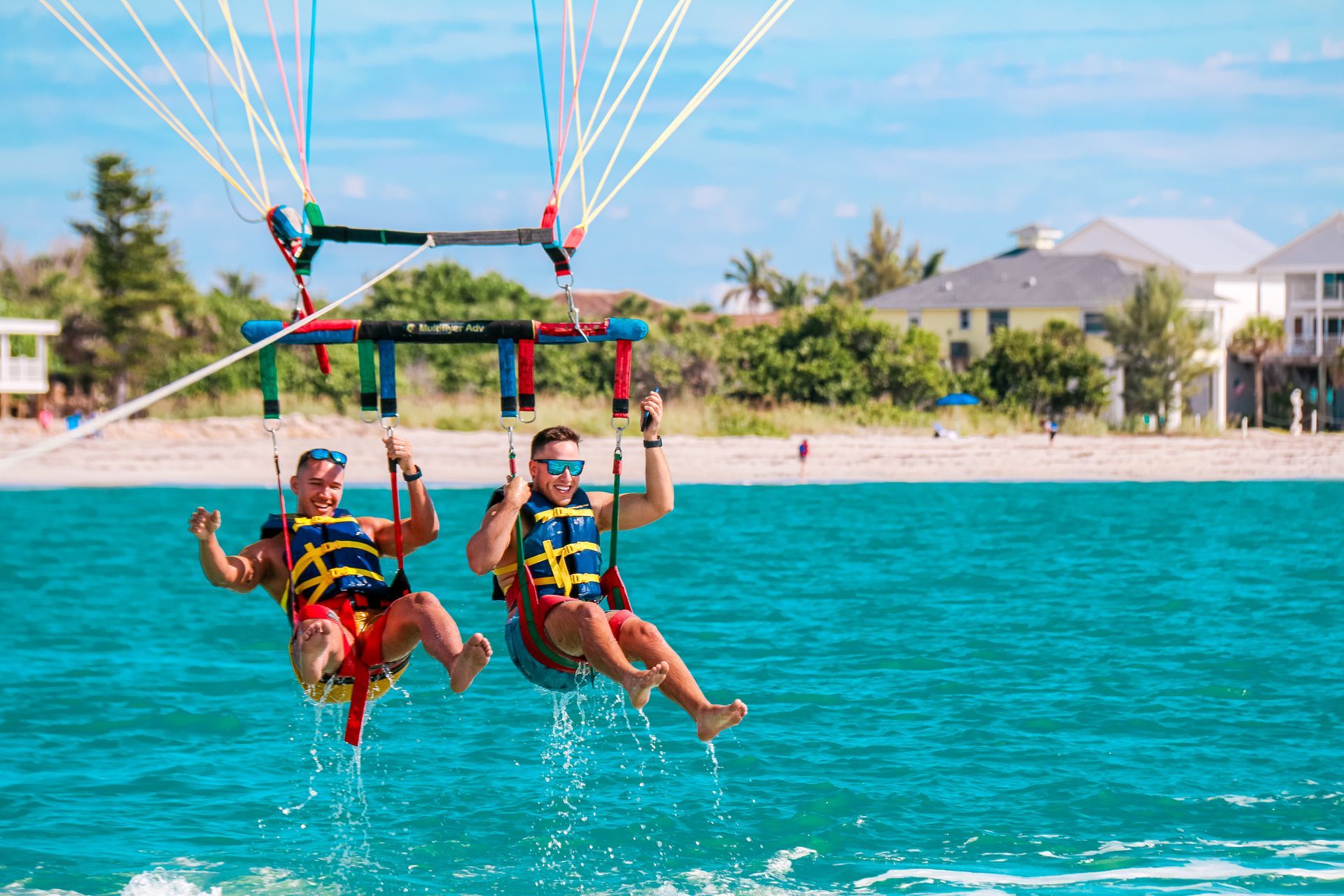 Two people parasailing over turquoise water near a beach with buildings in the background.