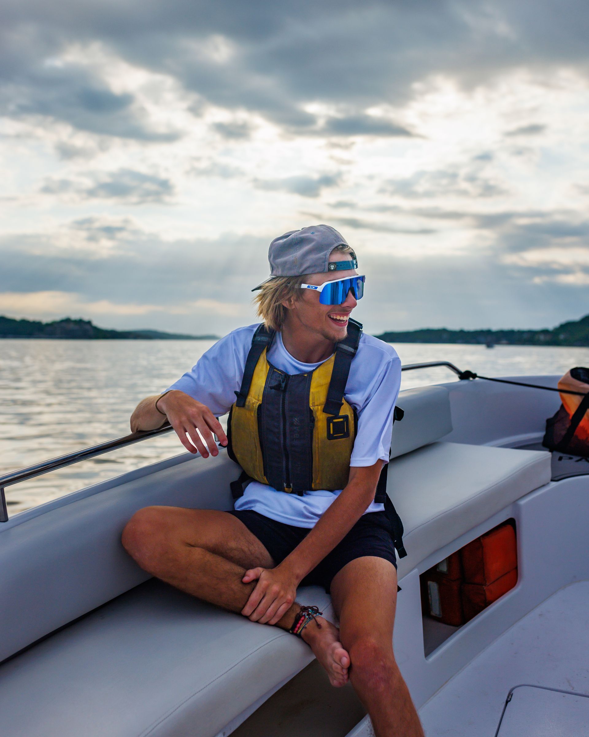 Young person on a boat wearing sunglasses and life vest, smiling at an overcast sky.