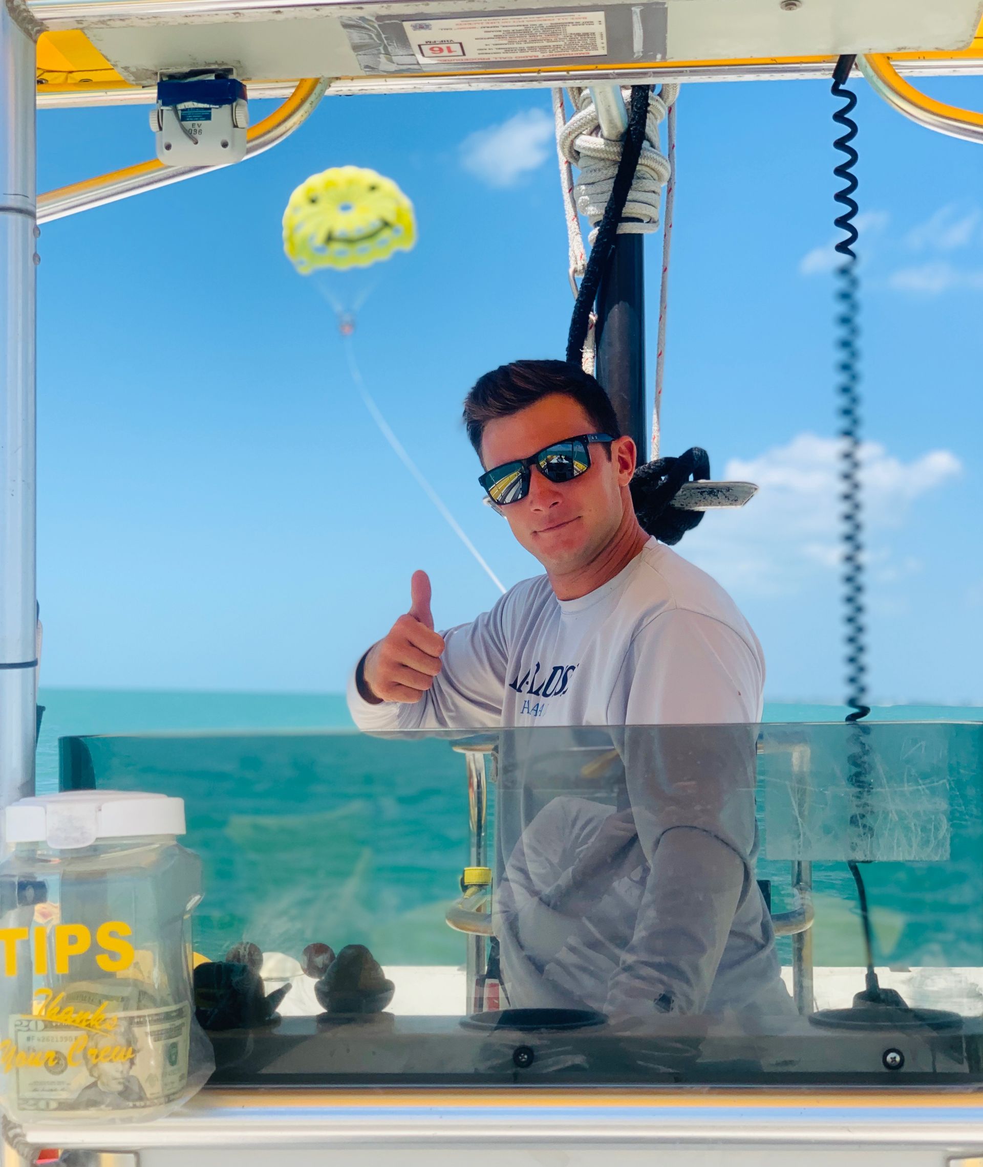 Man on boat gives thumbs up, with a smiley face balloon in the sky. Ocean in background.