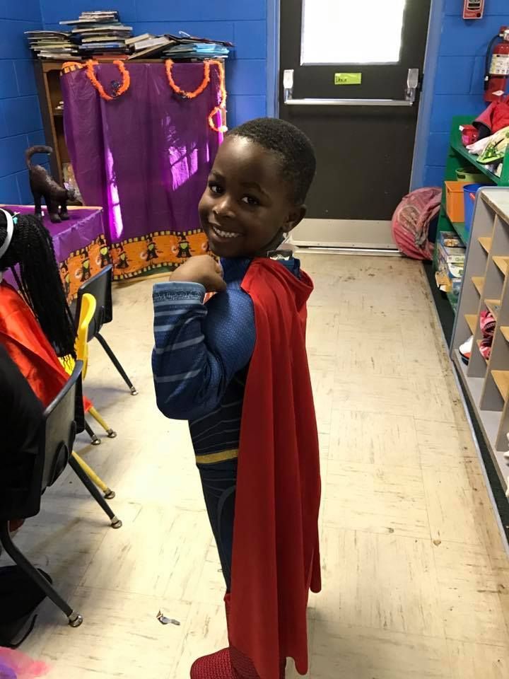 A young boy in a superman costume is standing in a hallway.