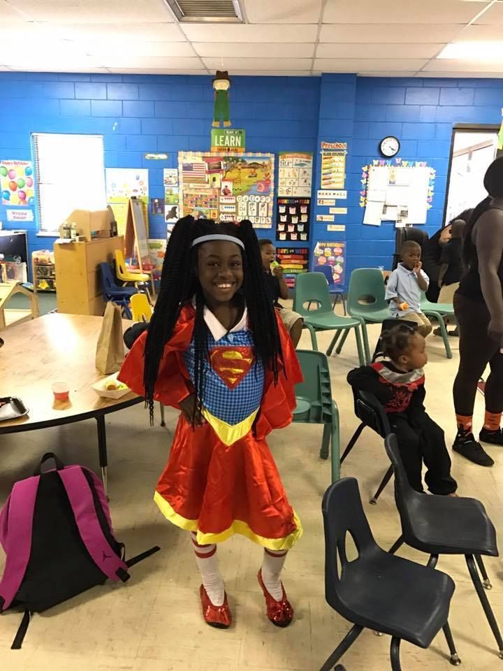 A young girl in a superman costume is standing in a classroom.