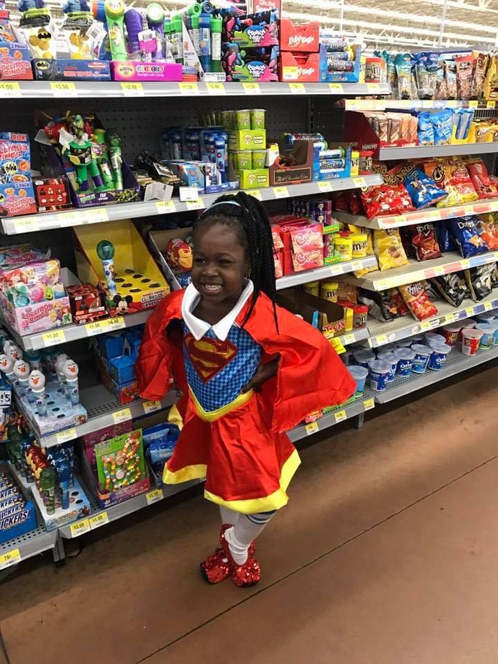 A little girl in a superman costume is standing in front of a shelf in a store.