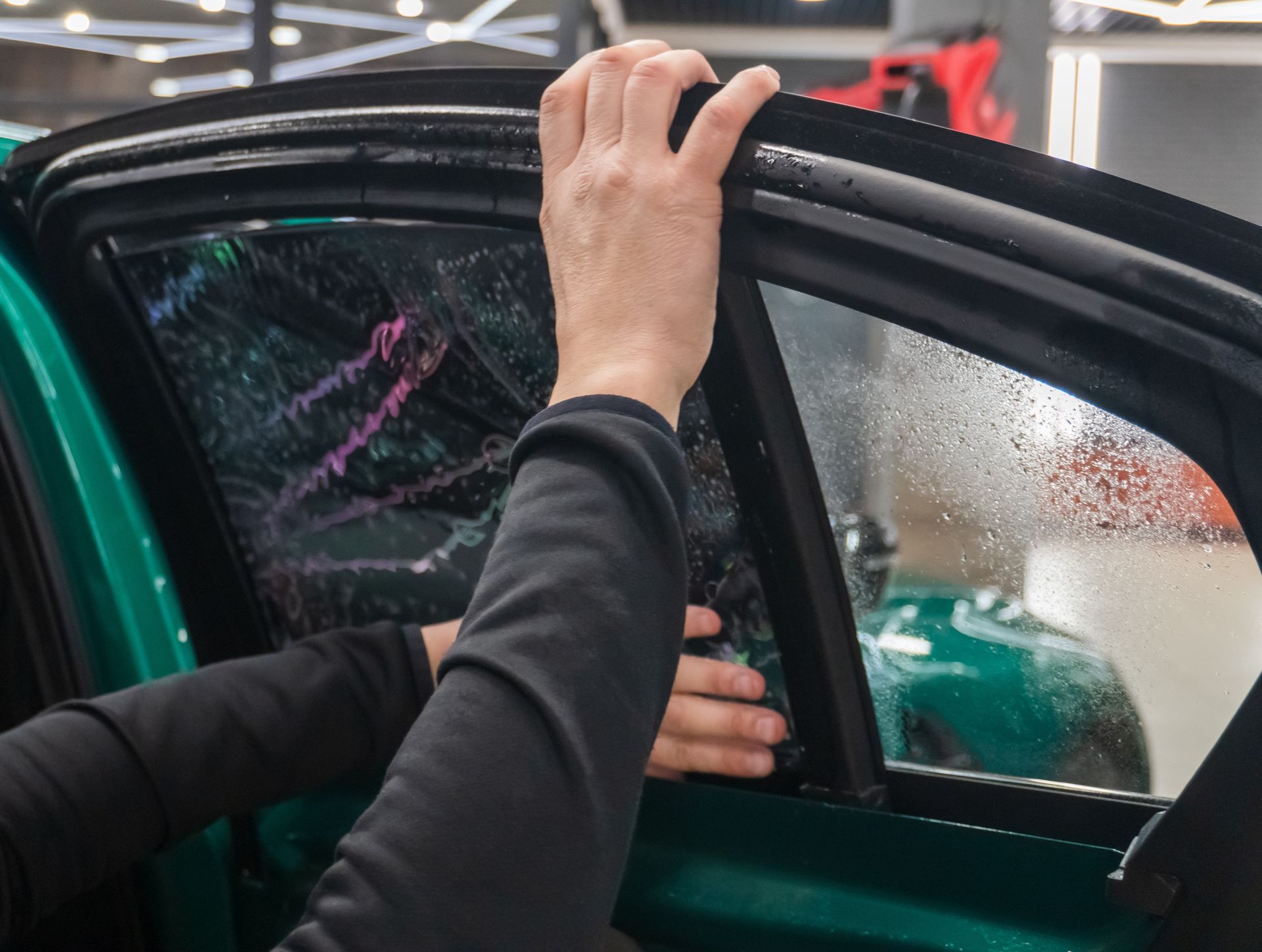 Hand adjusting a car door frame beside a green vehicle, with rain-speckled glass in a showroom