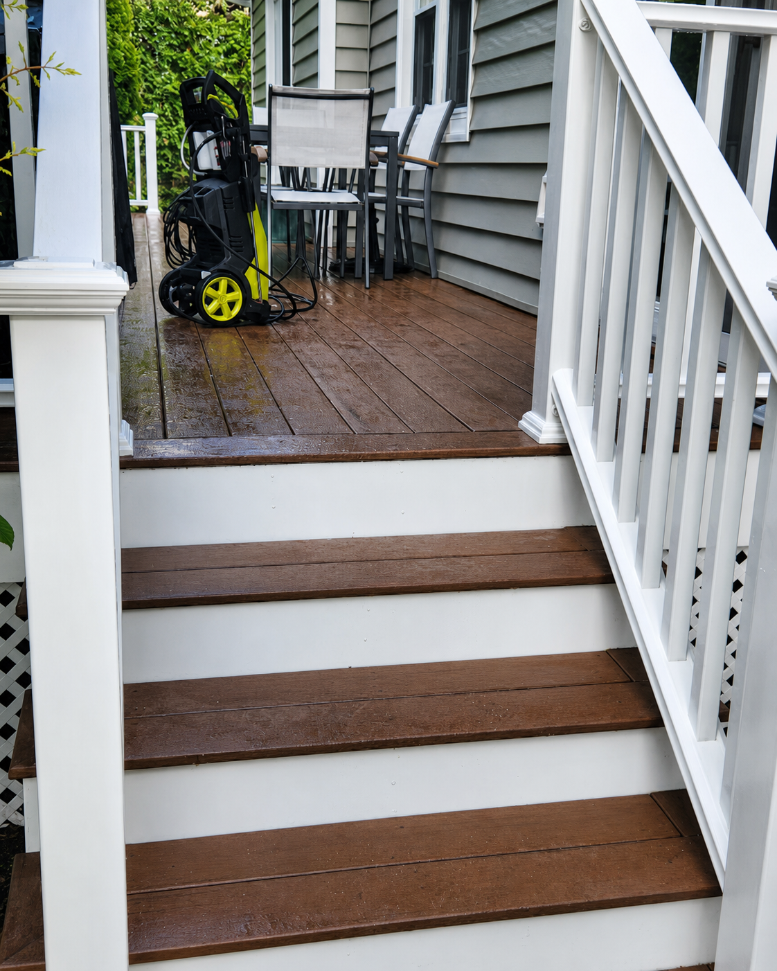 Front porch steps with white railings, brown treads, and a pressure washer on the wet deck landing.