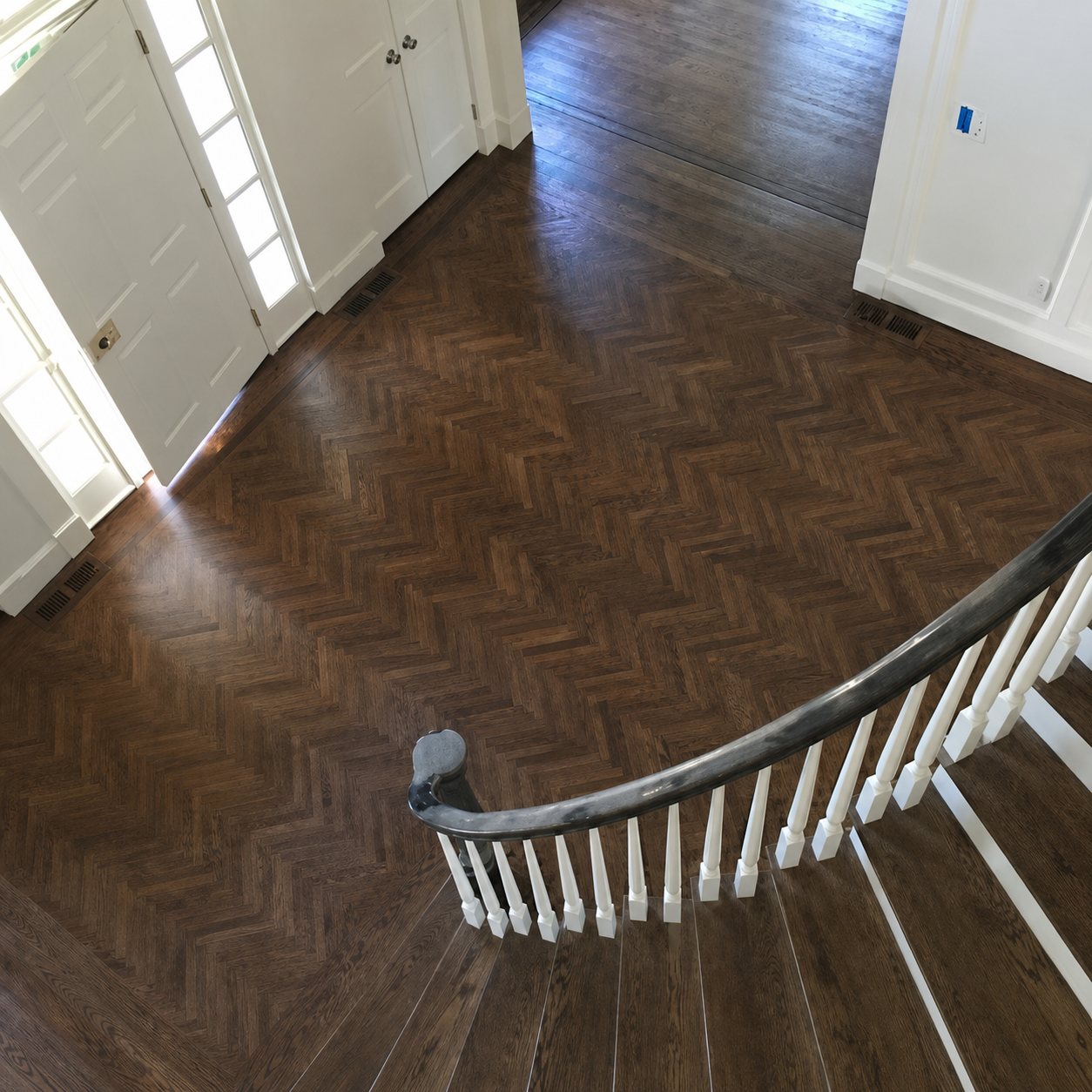 Curved staircase overlooking a dark herringbone wood floor in a bright hallway